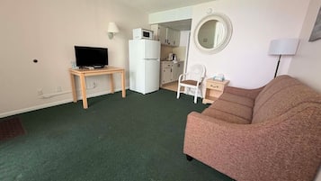 A compact living space with a sofa, a television on a small table, a kitchenette, and a mirror on the wall.