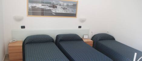 A hotel room with two single beds, a small bedside table with a lamp, a wall-mounted picture, and a plain white wall.