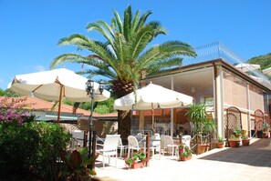 An outdoor patio area with white chairs and tables, umbrellas, and potted plants. - Hotel Villa Wanda (Porto Azzurro)