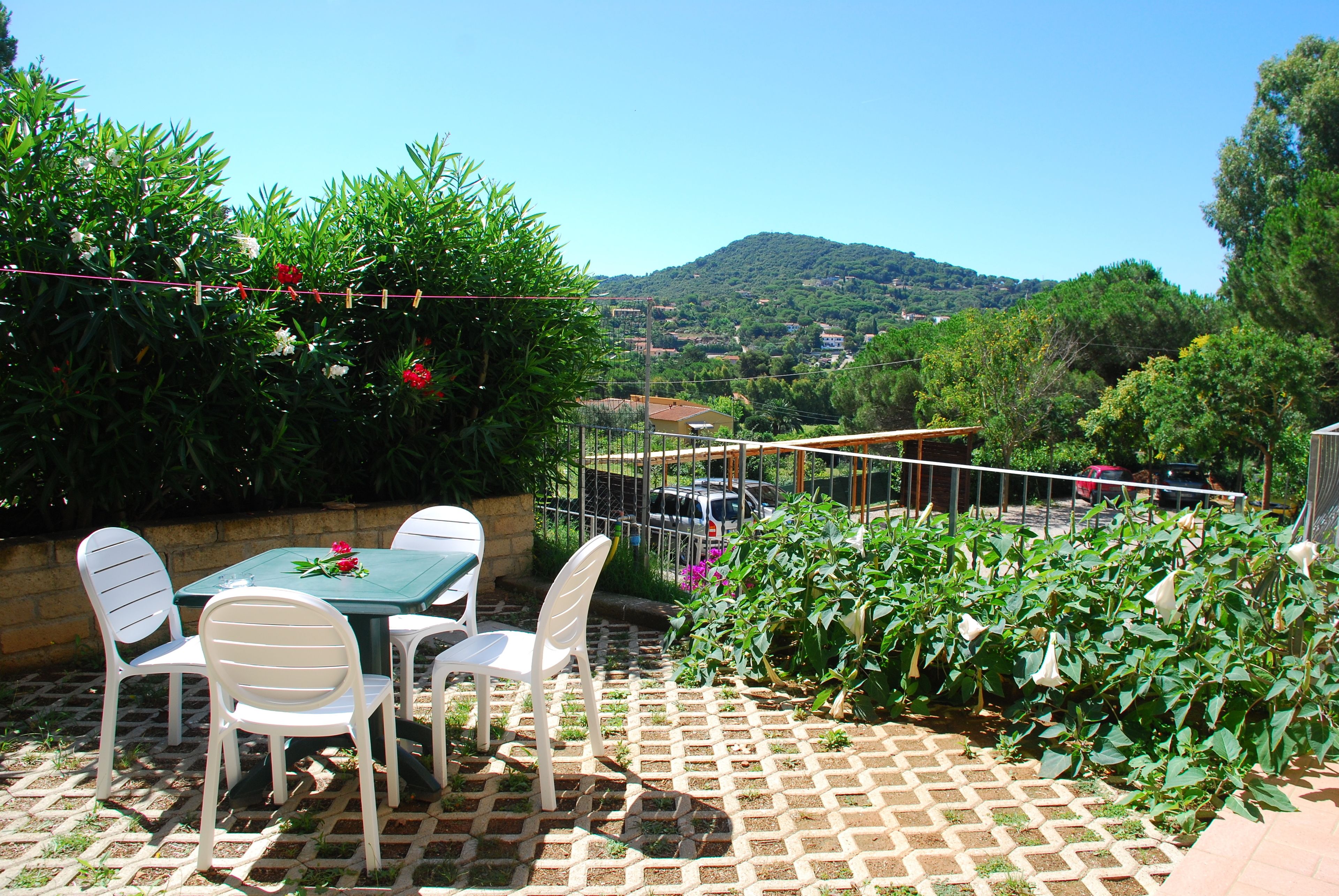 An outdoor patio with a table and chairs, surrounded by lush greenery and a view of a hill in the distance.