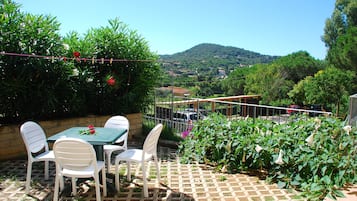 An outdoor patio with a table and chairs, surrounded by lush greenery and a view of a hill in the distance.