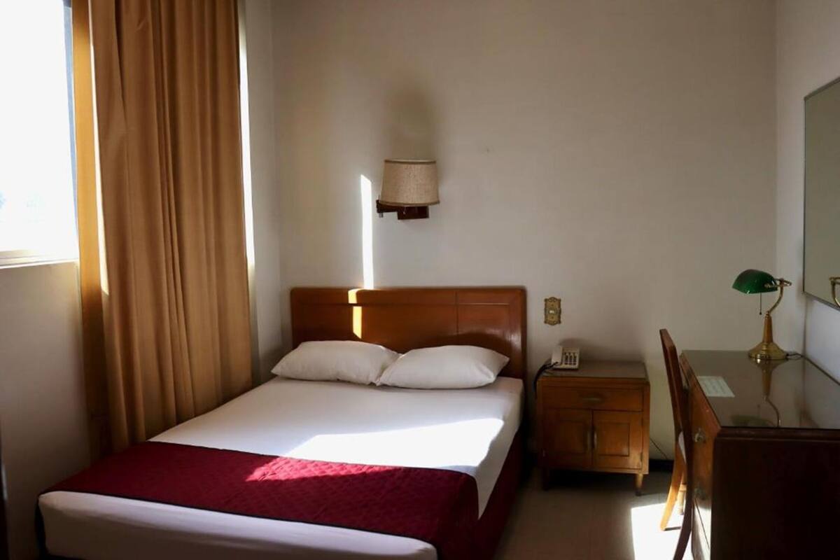 A hotel room with a bed, a desk, a chair, a lamp, and a wall-mounted light fixture.