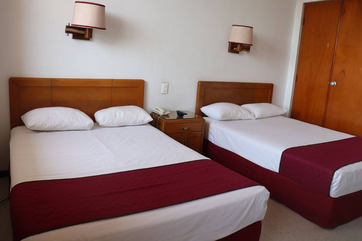 A hotel room with two single beds, each with a wooden headboard, white bedding, and a red accent at the foot of the bed. There are bedside tables with lamps and a telephone.