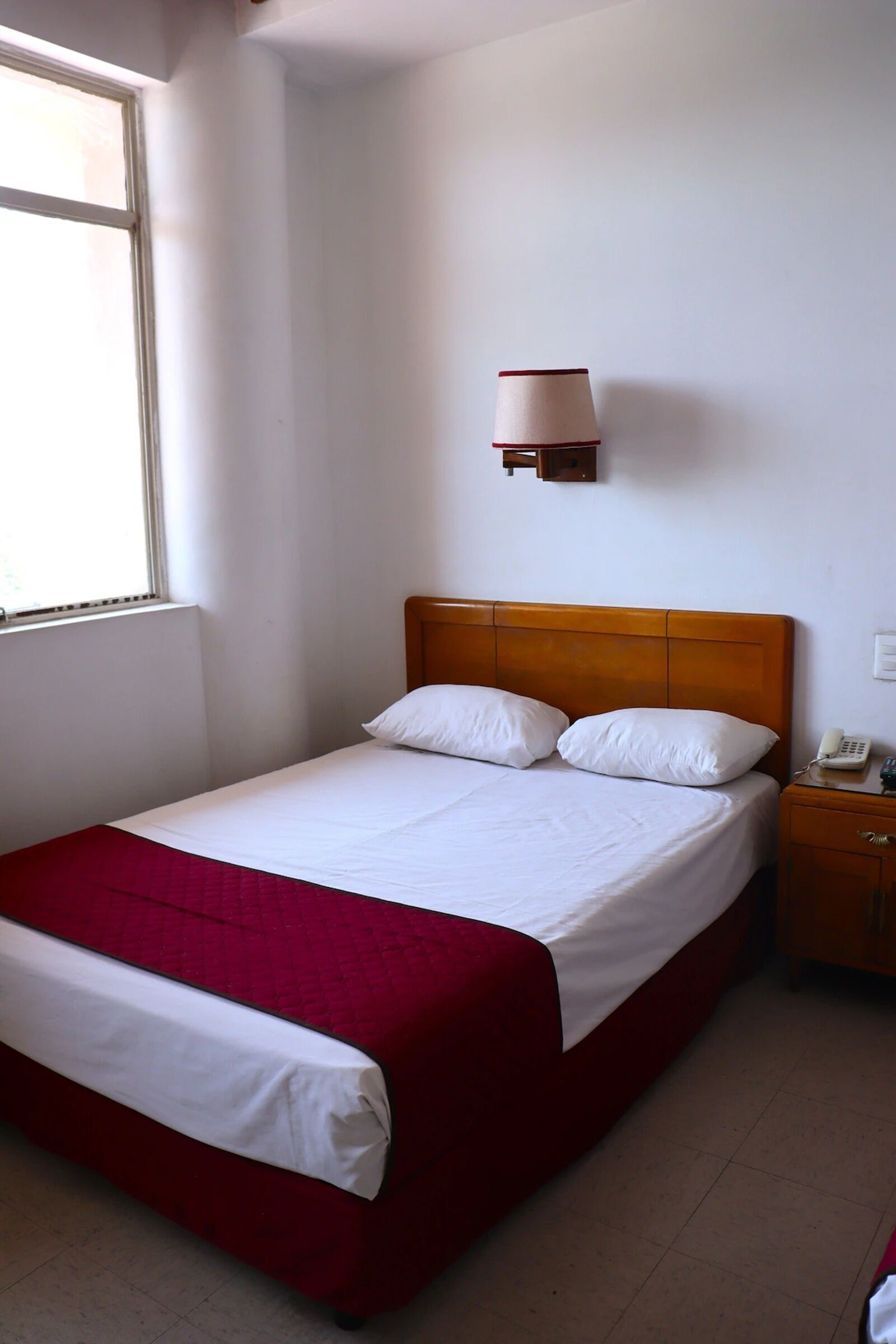 Una cama individual con cabecera de madera, una mesita de noche con teléfono y una ventana que deja entrar luz natural.