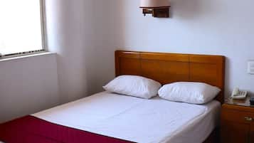 Una cama individual con cabecera de madera, una mesita de noche con teléfono y una ventana que deja entrar luz natural.