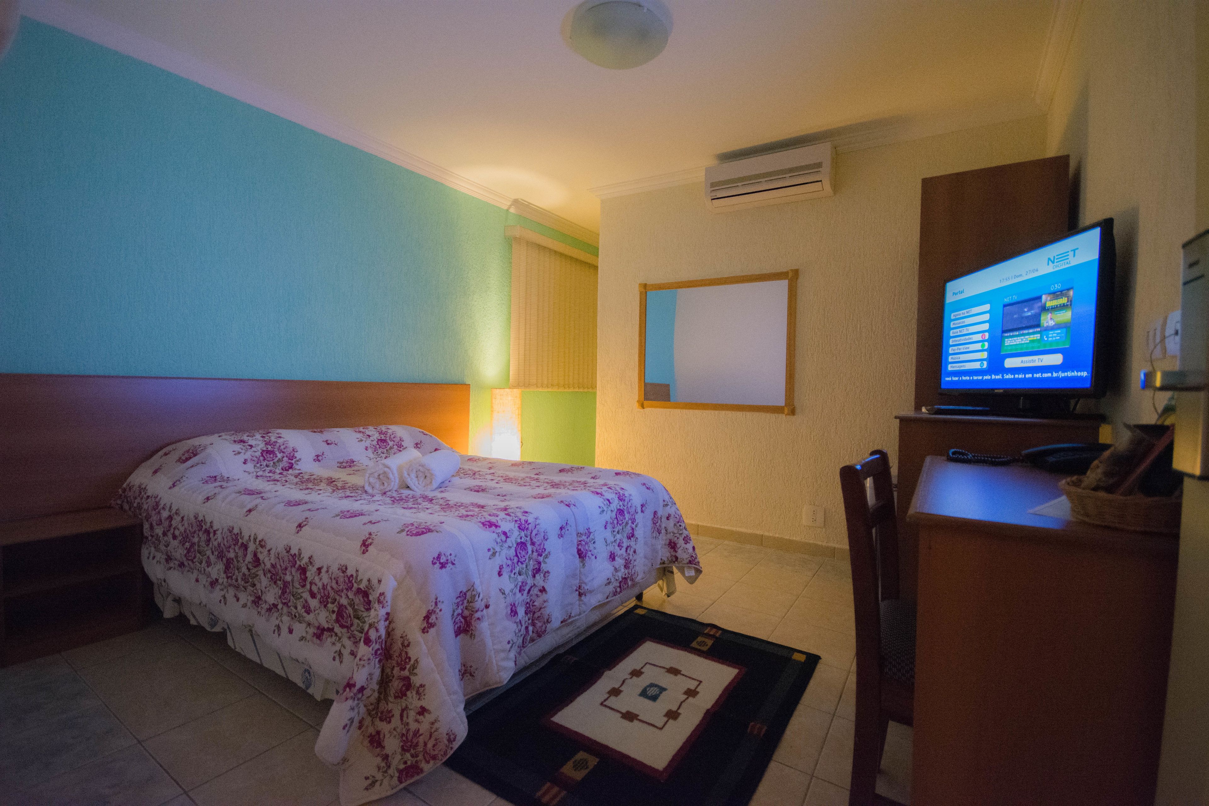 Quarto de hotel com cama, escrivaninha, cadeira, televisão e ar condicionado.