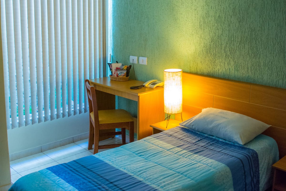 A hotel room with a bed, a desk with a lamp, a chair, and a window with blinds.