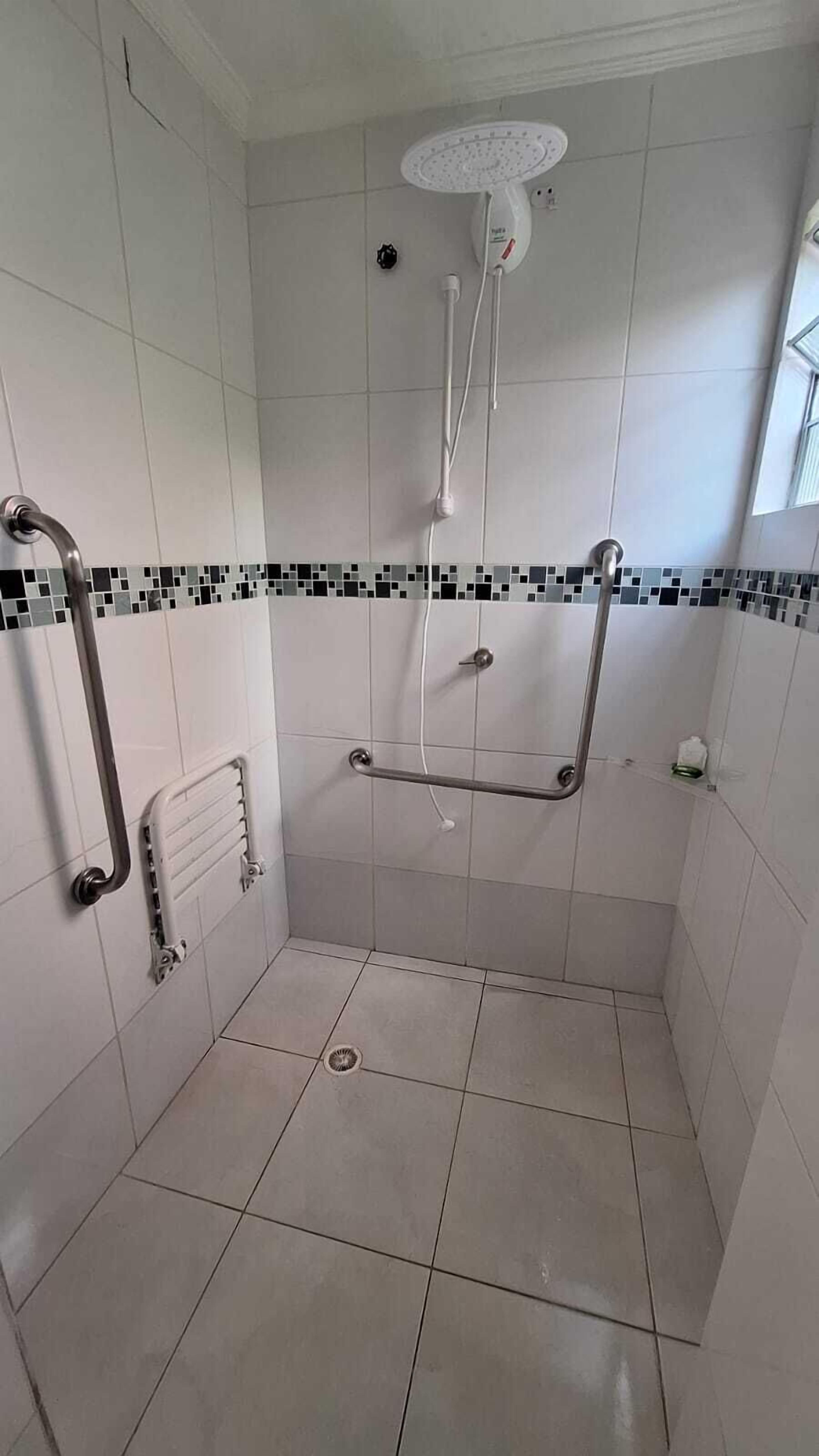 Área de chuveiro com azulejos brancos, chuveiro manual e barra de apoio.