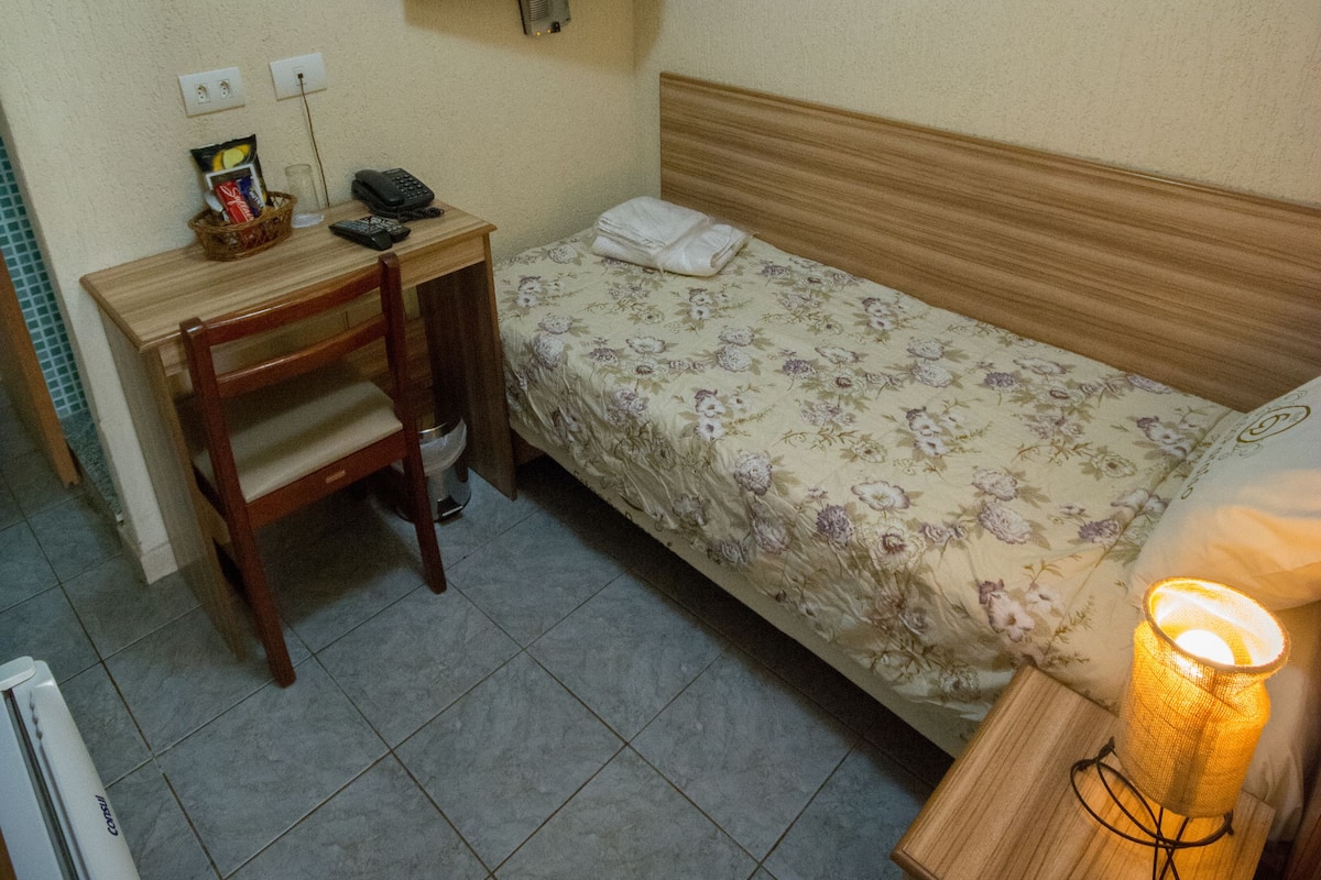 A small, neatly arranged room with a bed, a desk, a chair, a television mounted on the wall, and a bedside lamp.