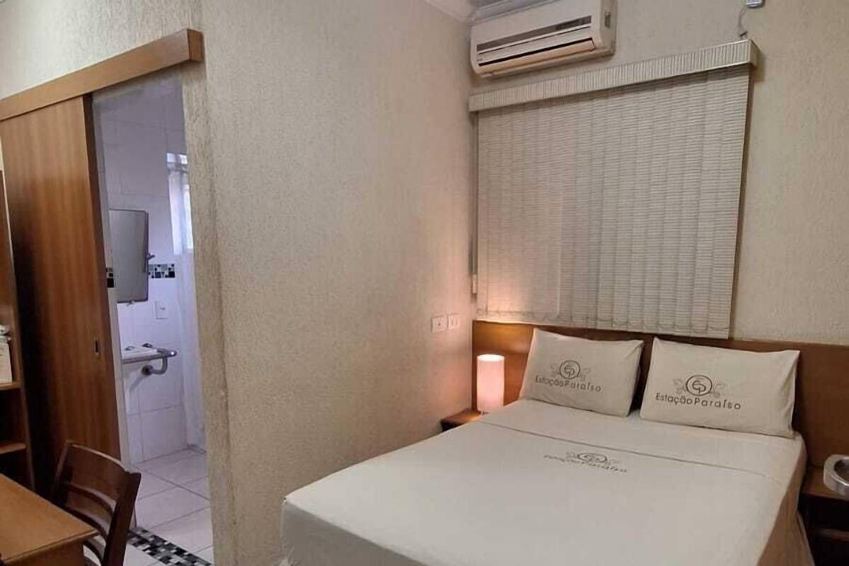 A hotel room with a bed, a desk, an air conditioning unit, and a bathroom visible through an open door.