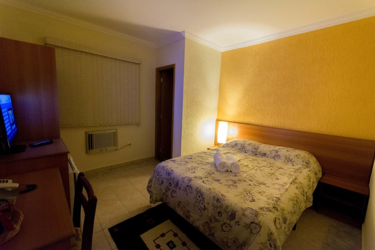 A hotel room with a bed, a desk with a computer, a chair, a window with blinds, and a wall-mounted lamp.