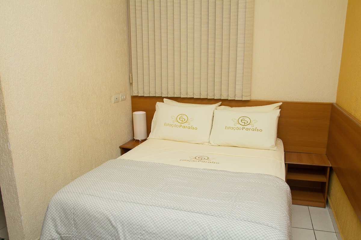 A hotel room with a bed, two pillows with the hotel's logo, a bedside table, and a window with blinds.