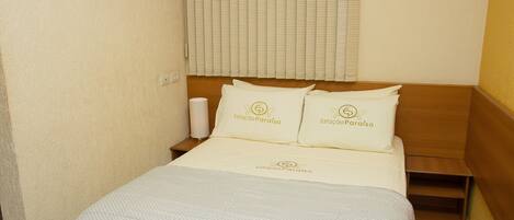 A hotel room with a bed, two pillows with the hotel's logo, a bedside table, and a window with blinds.