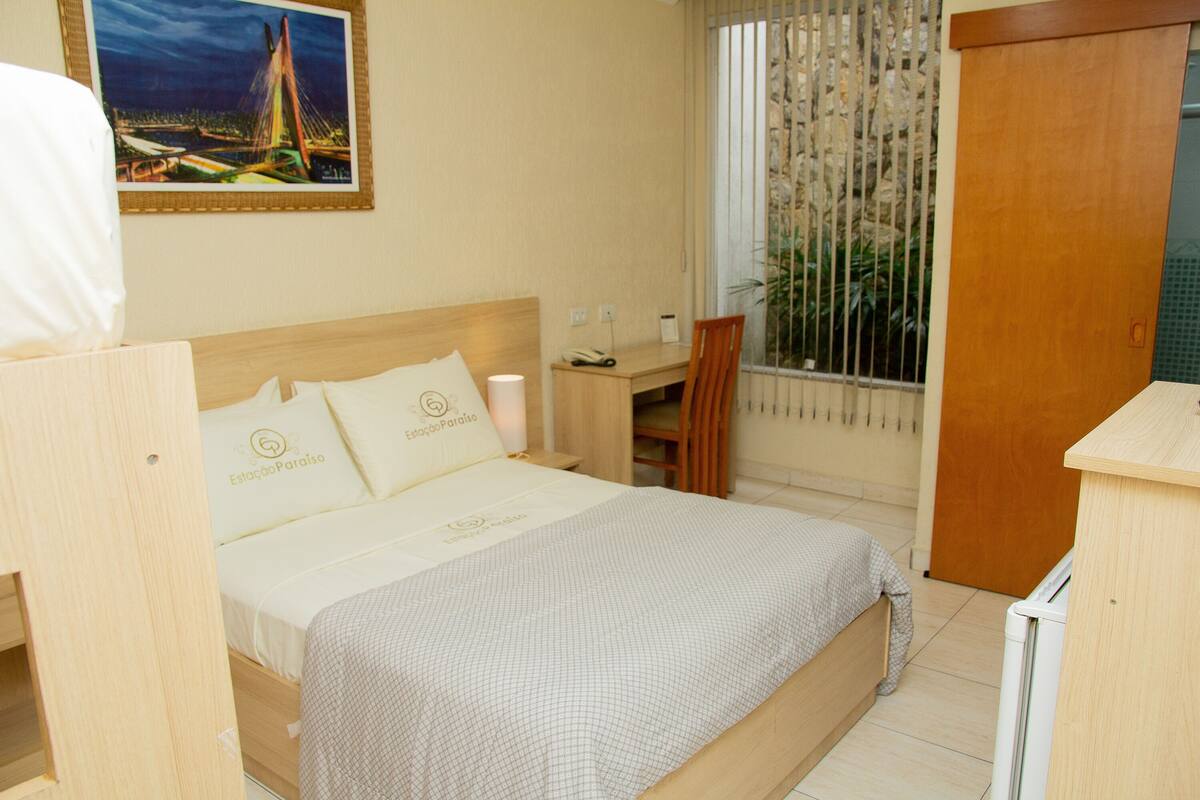 A hotel room with a bed, a desk, a chair, a window with blinds, and a wall-mounted picture.