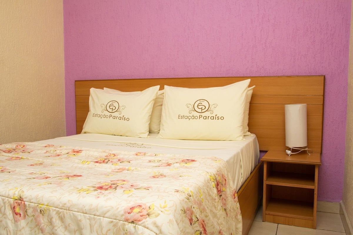 A hotel room with a bed, two pillows with the hotel's logo, a bedside table with a lamp, and a floral bedspread.