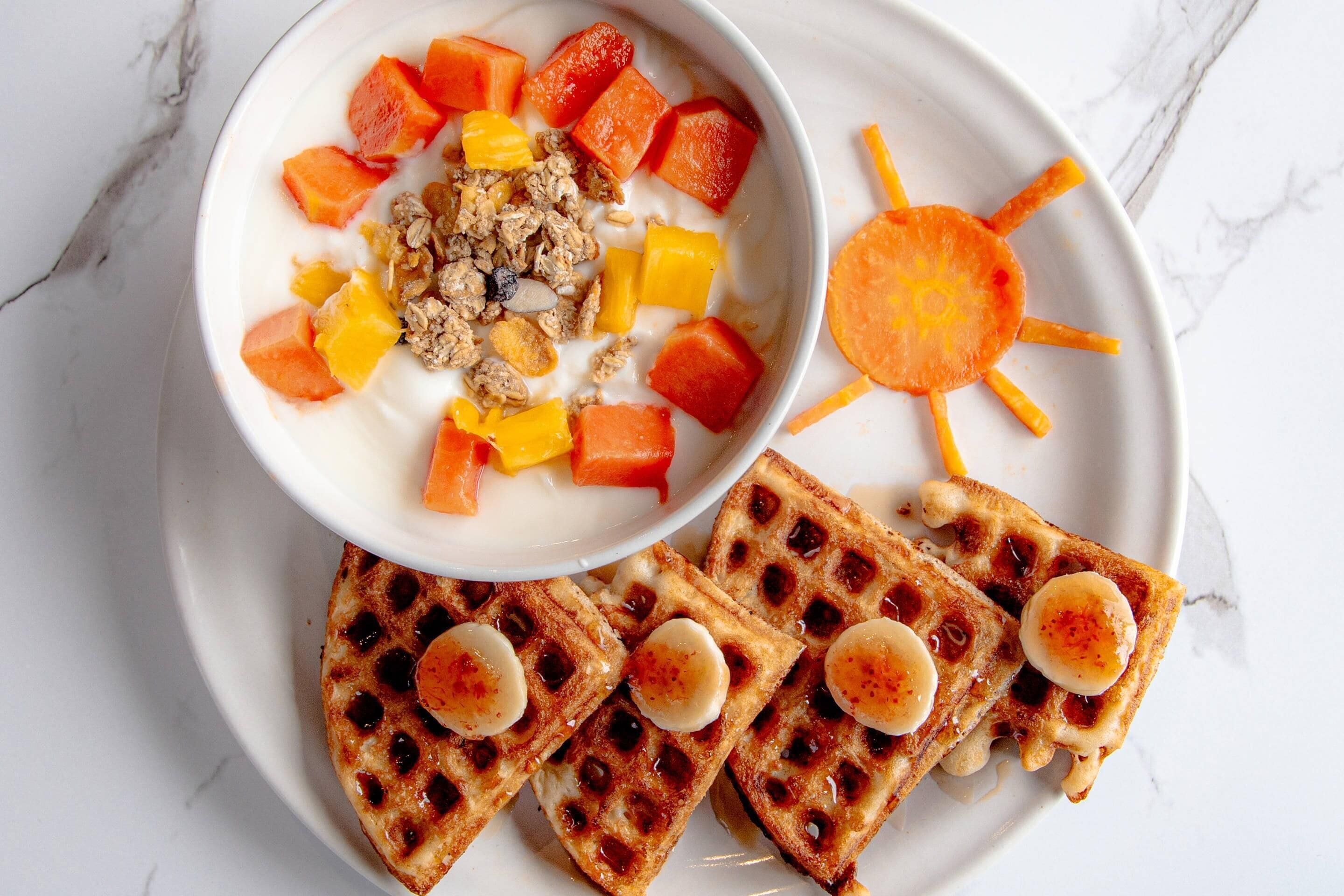 A plate with waffles topped with bananas and syrup, a bowl of yogurt with fruit and granola, and a sun-shaped carrot.