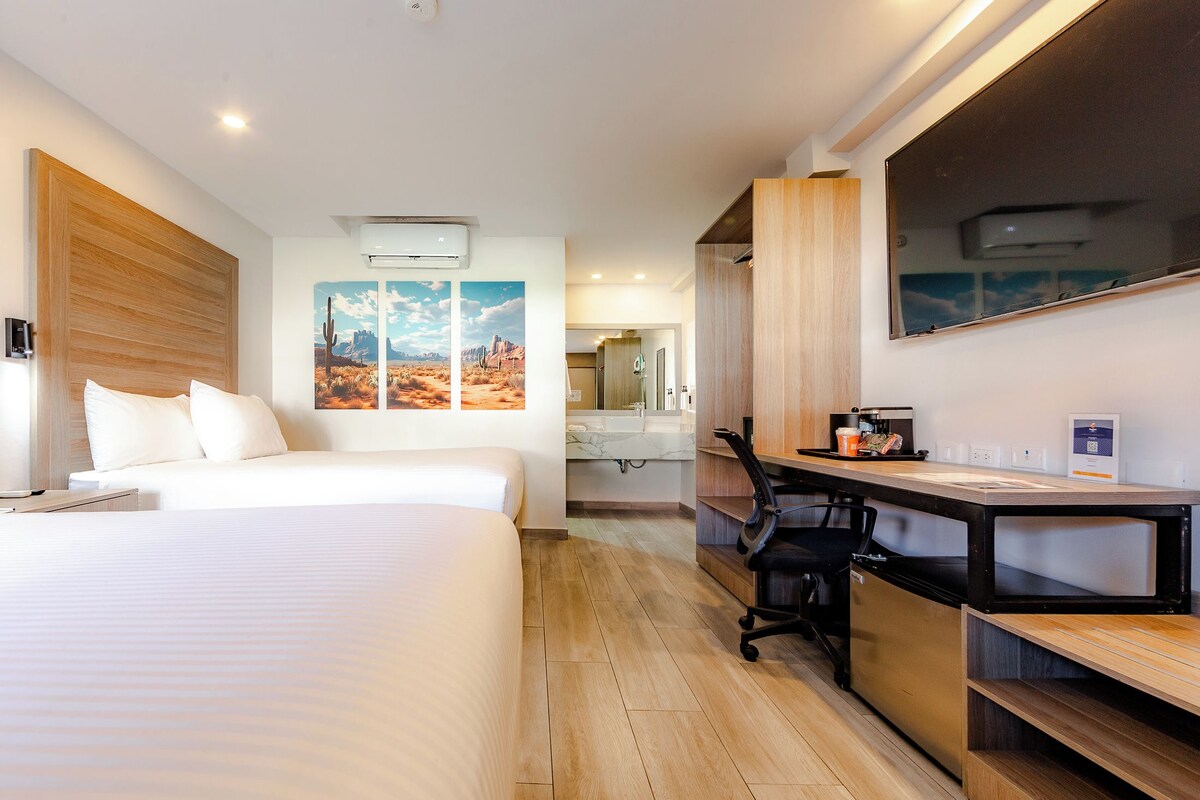 A hotel room with a bed, desk, chair, and artwork on the wall.