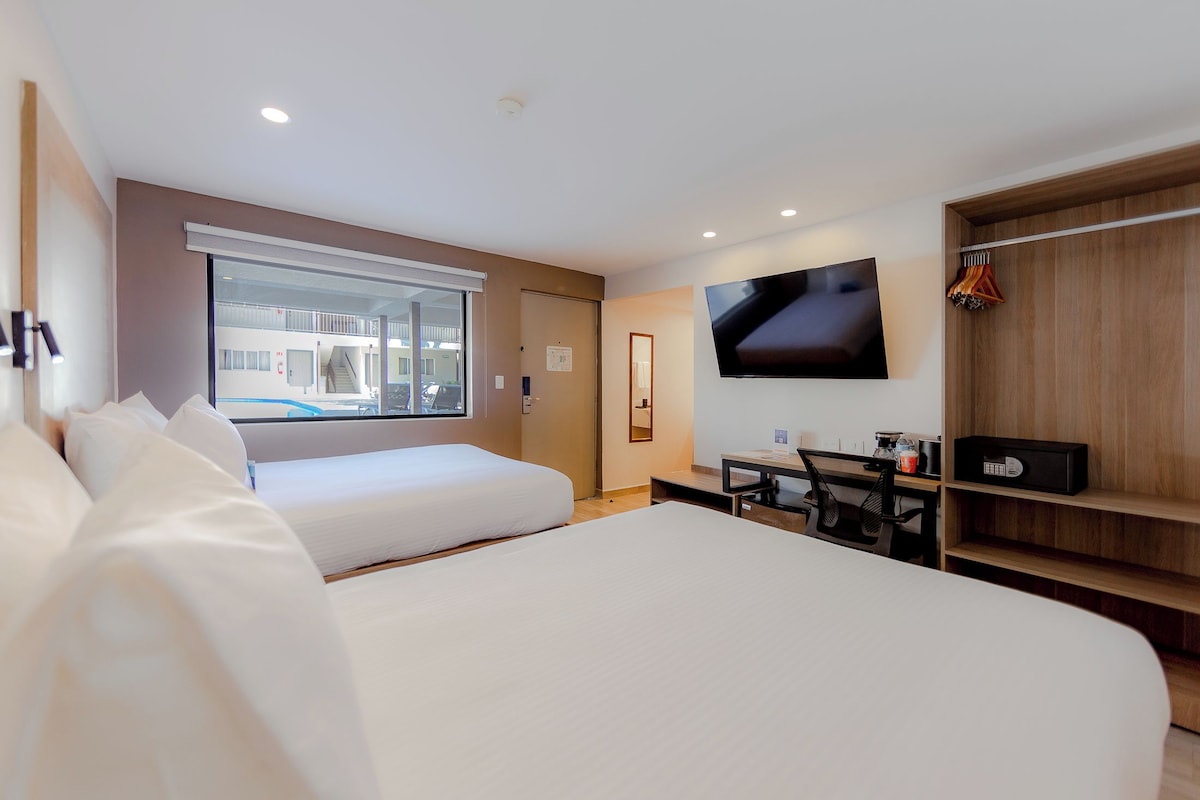 A hotel room with a large bed, a desk with a chair, a TV mounted on the wall, and a window offering a view of the outside.