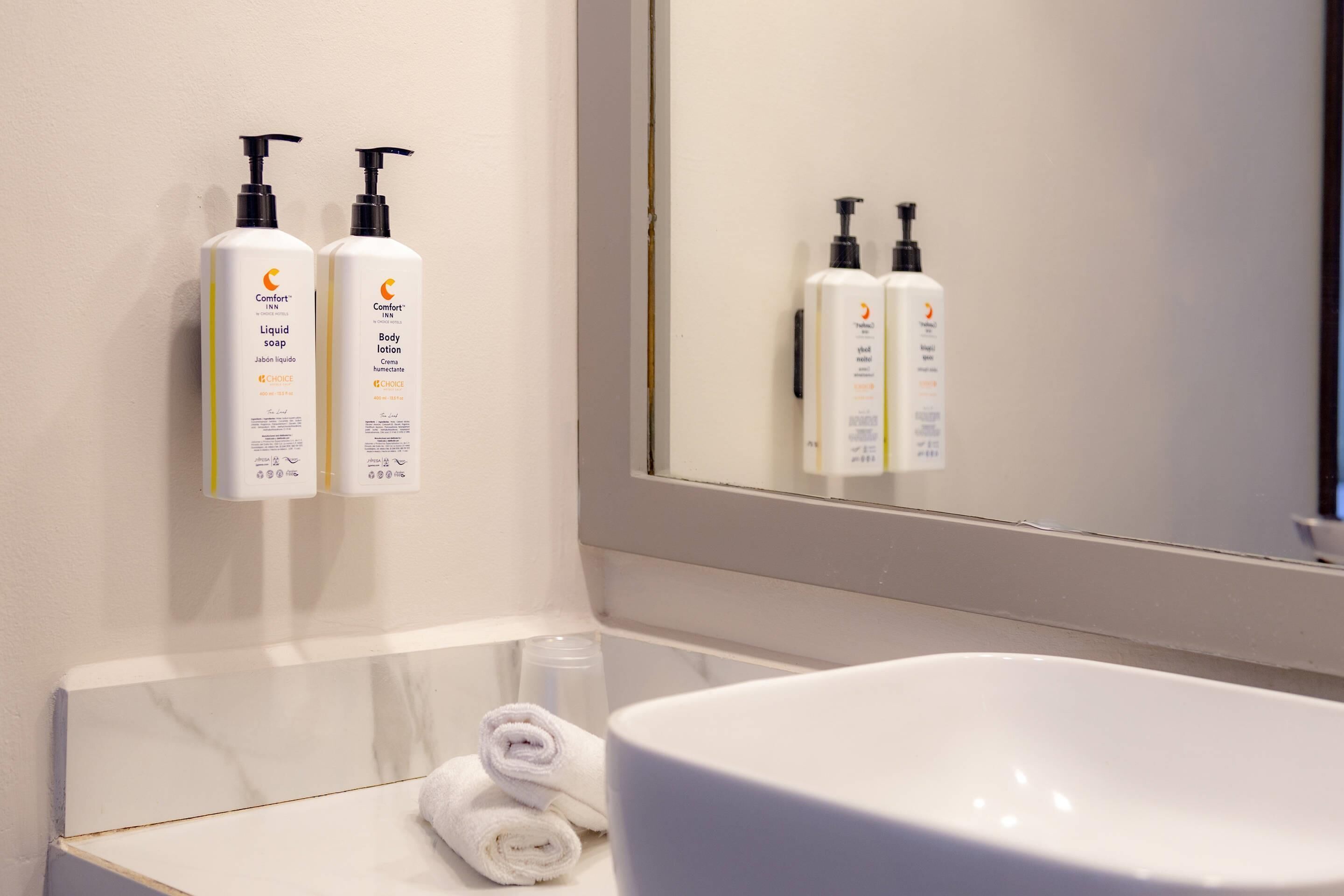 Un baño con un lavamanos blanco, dos botellas de loción corporal y un espejo que refleja las botellas.