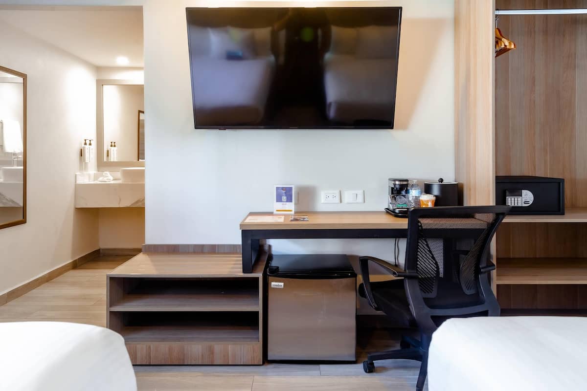 Habitación de hotel con cama, escritorio, computadora, televisor, mini refrigerador y una silla.