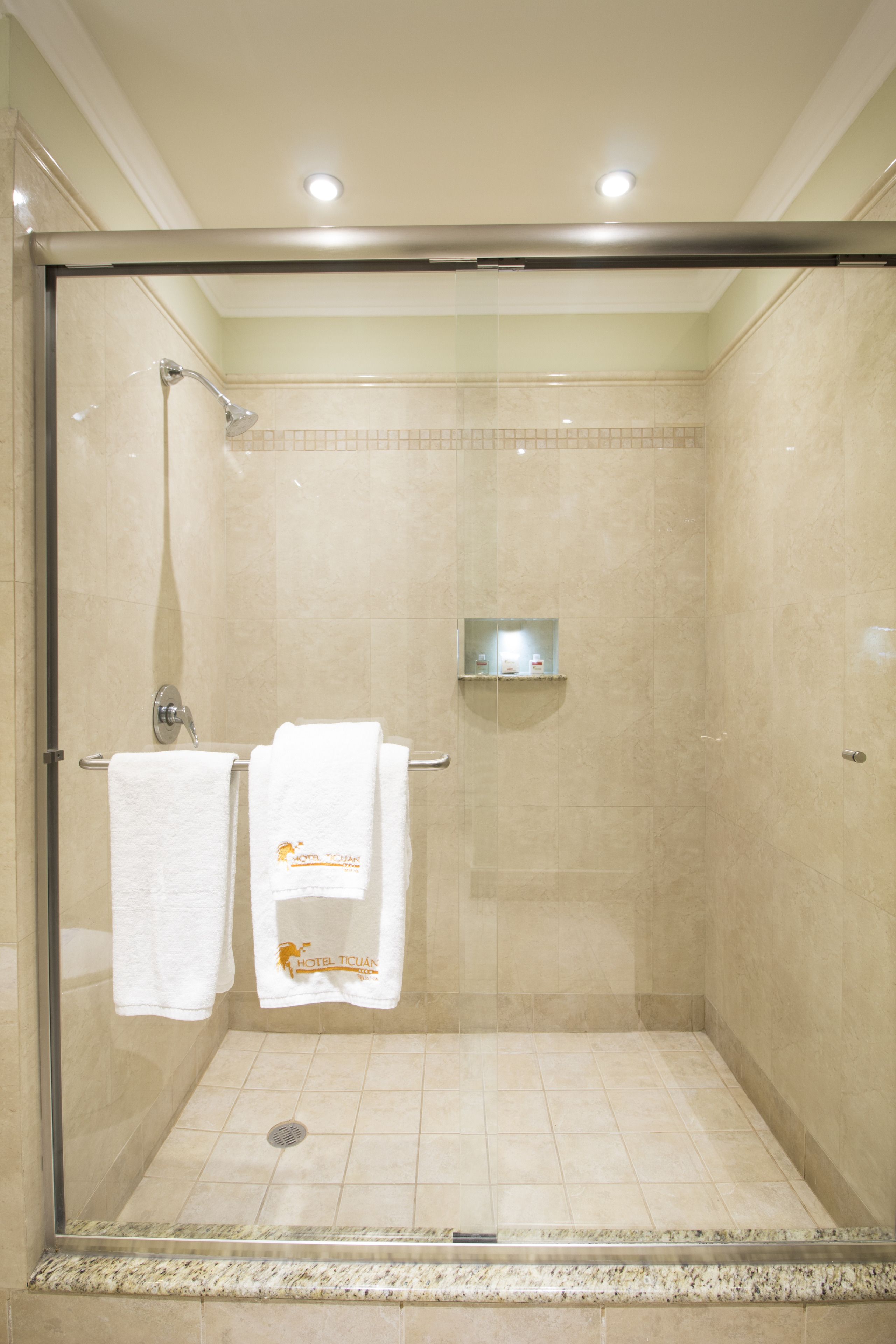 A glass-enclosed shower with a metal frame, beige tiled walls, and a small shelf with toiletries.