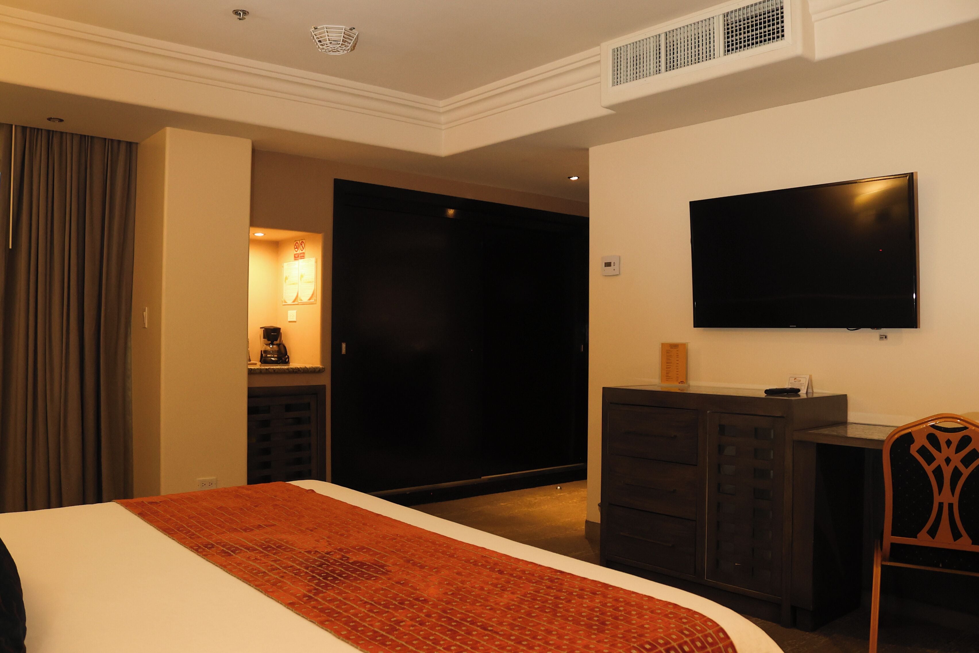 Habitación de hotel con una cama, un televisor montado en la pared, un aparador y una puerta que da a otra habitación.