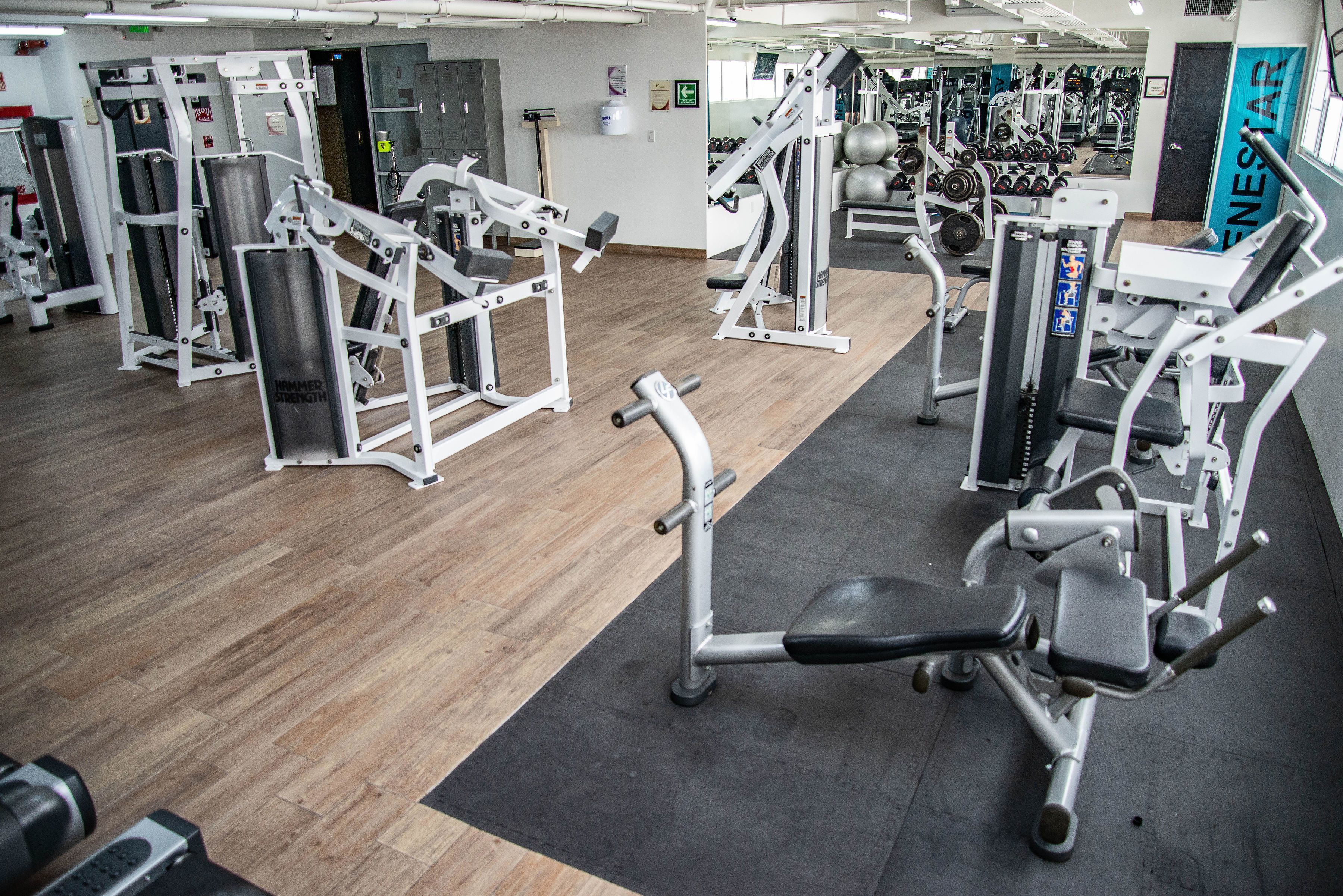 Un gimnasio con diversas máquinas de ejercicio, suelo de madera y una colchoneta negra en el centro.
