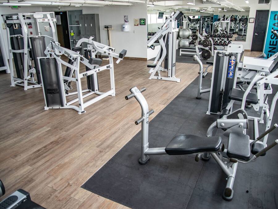 Un gimnasio con diversas máquinas de ejercicio, suelo de madera y una colchoneta negra en el centro.