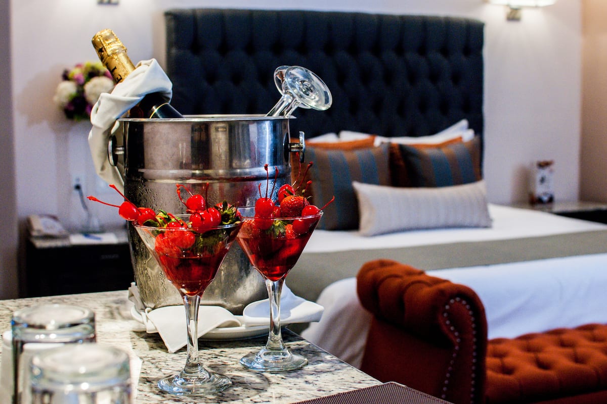 Un mini-bar avec une bouteille de champagne et deux verres à martini remplis d’une boisson rouge, décorés de cerises, sur une table en marbre dans une chambre d’hôtel.