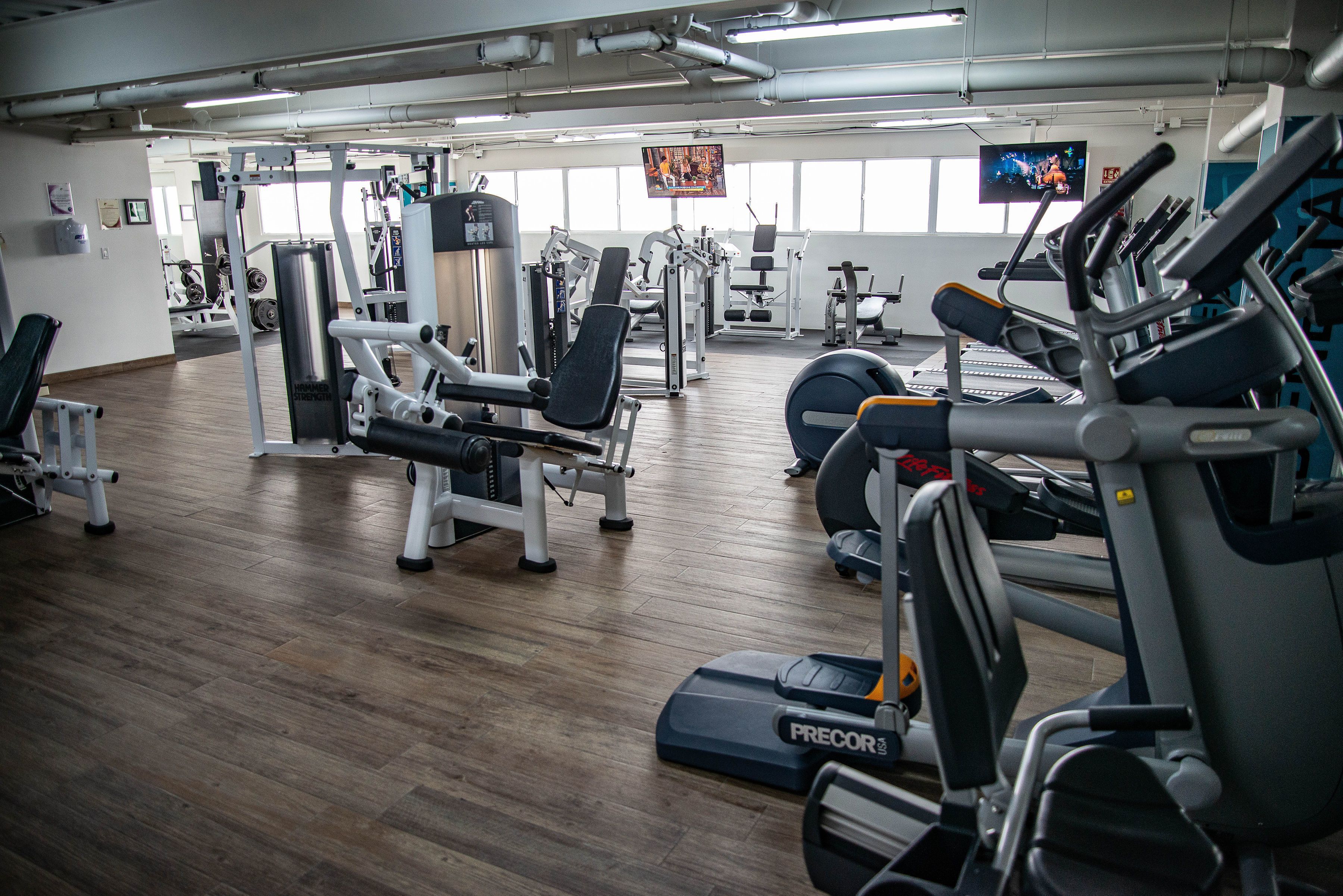 Een fitnessruimte met diverse fitnessapparatuur, waaronder loopbanden en krachtapparaten, en een houten vloer.