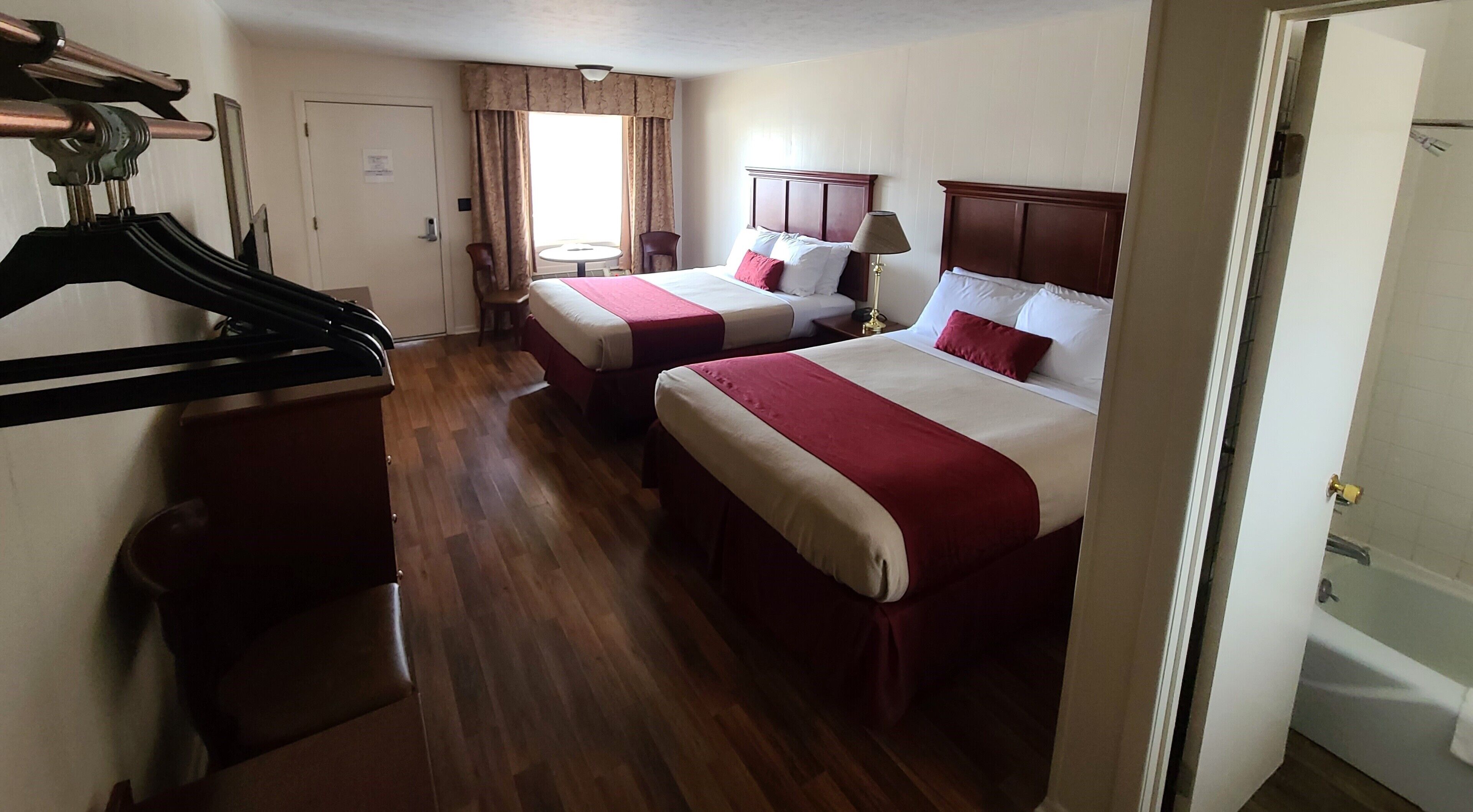 A hotel room with two beds, wooden flooring, a coat rack, a chair, and a window with curtains.