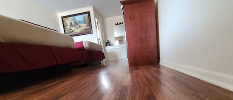 A room with wooden flooring, a bed with a red cover, a painting on the wall, and a wooden cabinet.