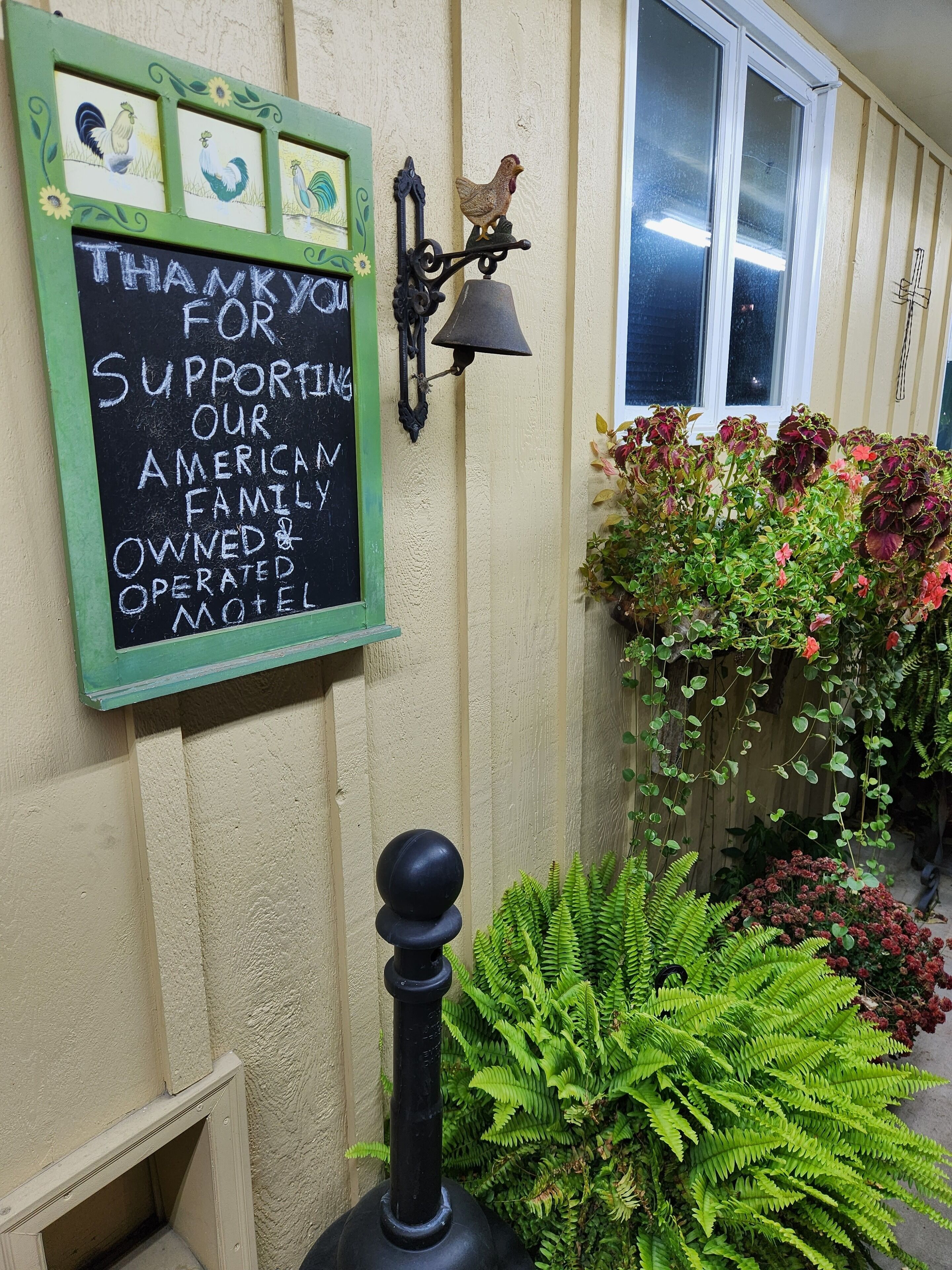 Un mur orné d’un panneau noir sur lequel est écrit un message de remerciement à l’intention des personnes qui soutiennent ce motel familial, ainsi qu’un coq décoratif et une végétation luxuriante.