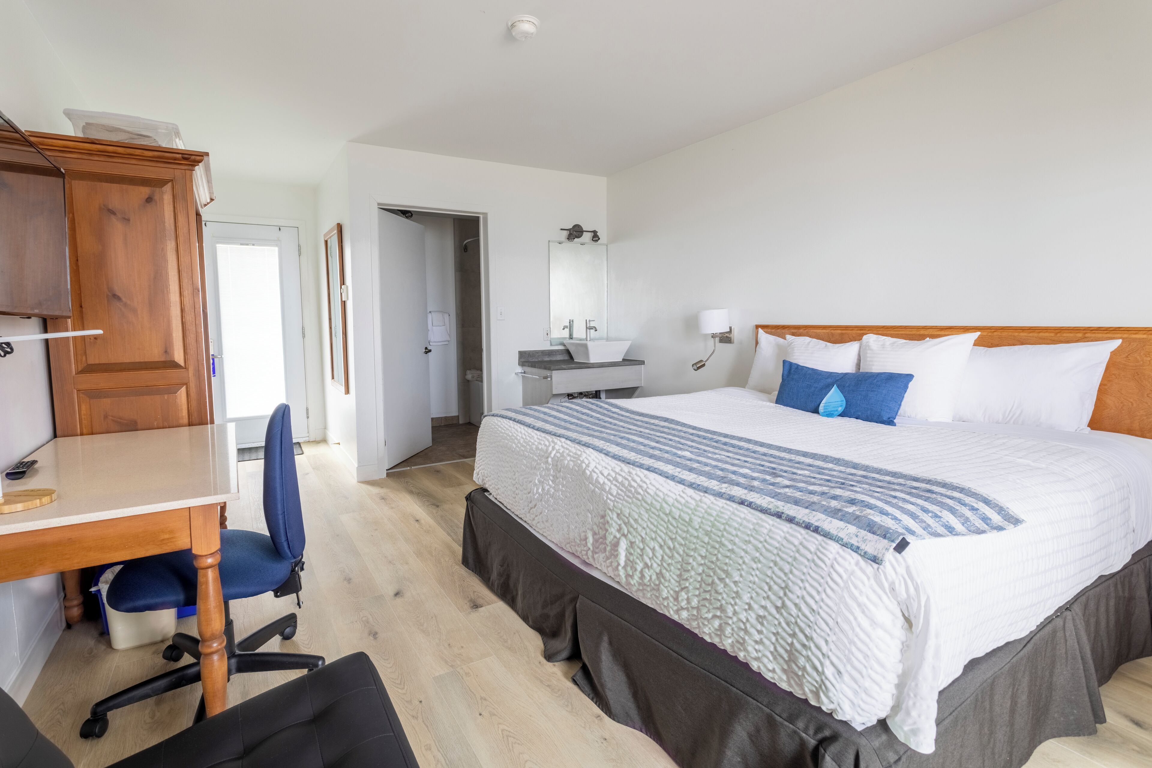 A hotel room with a wooden desk, a blue chair, a bed with white and blue bedding, a wooden headboard, a bathroom with a sink and mirror, and a door leading to another room.