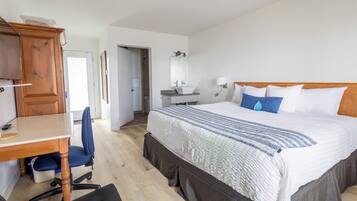 A hotel room with a wooden desk, a blue chair, a bed with white and blue bedding, a wooden headboard, a bathroom with a sink and mirror, and a door leading to another room.