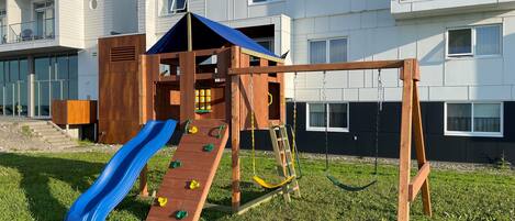 A playground with a slide and climbing wall, set against a modern building with multiple windows.