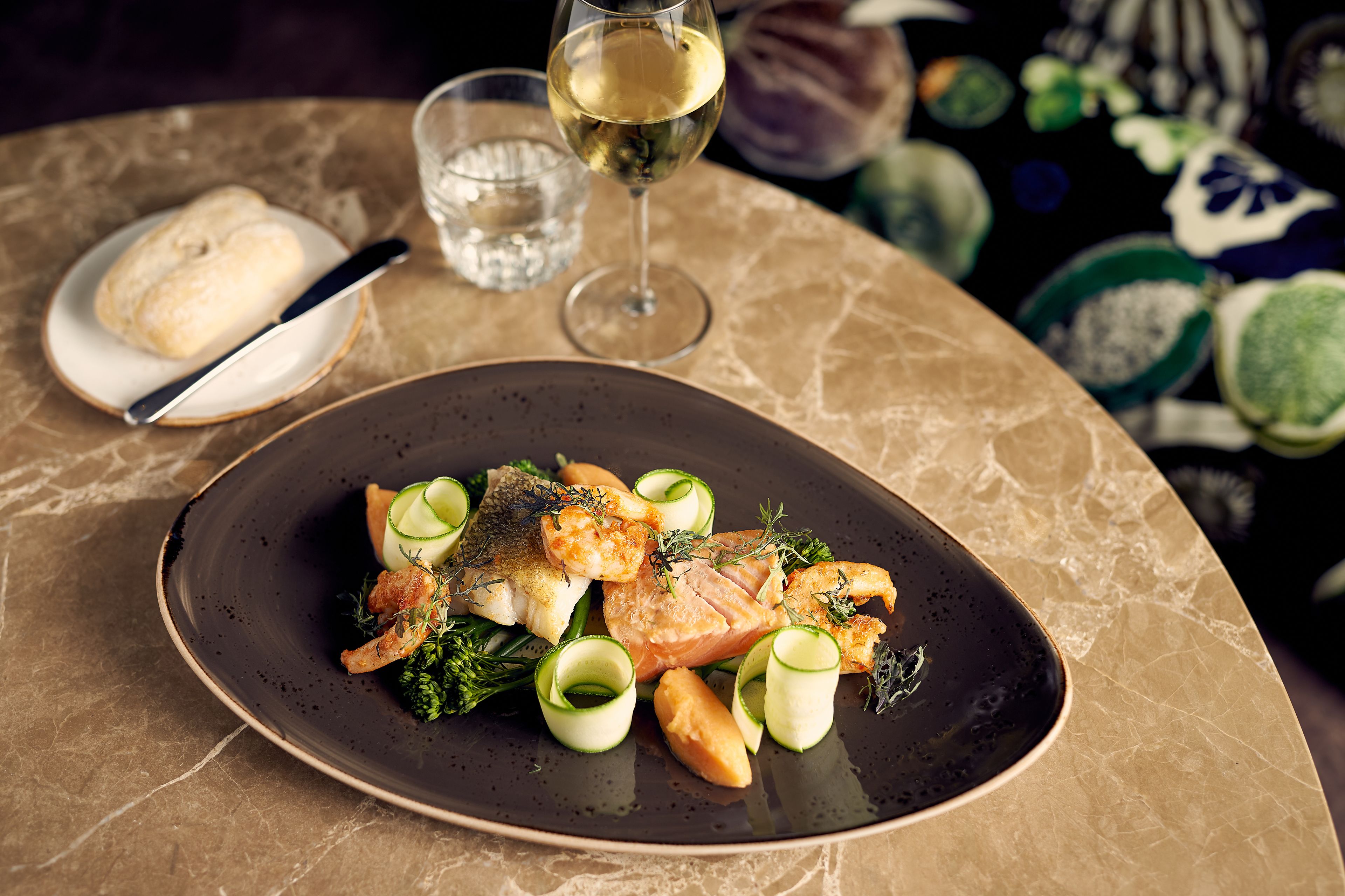 A plate of food with a glass of white wine and a small bowl of soup on a marble table.