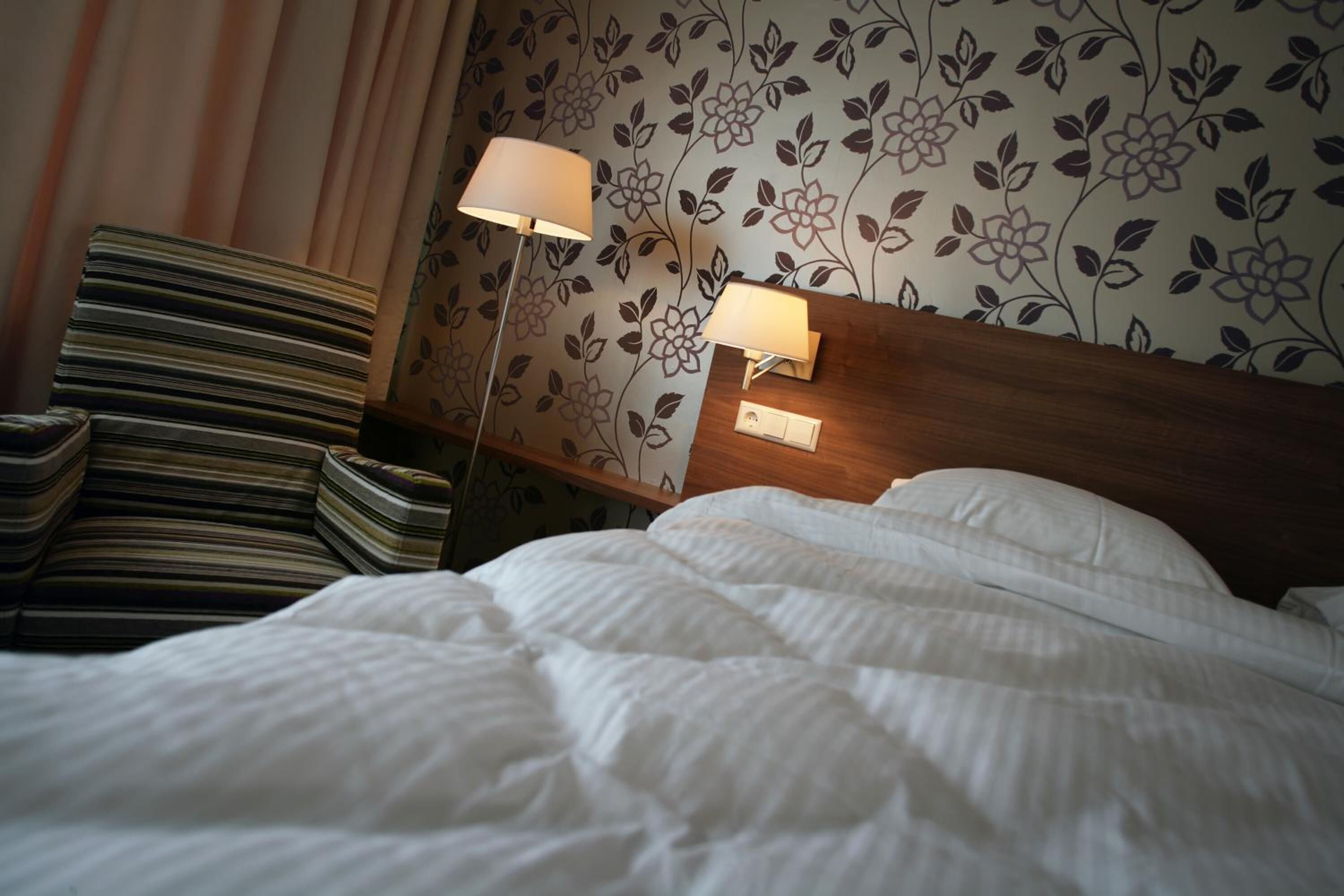 Een hotelkamer met een keurig opgemaakt bed, een stapel boeken, een gestreept fauteuil en een bloemen behang.