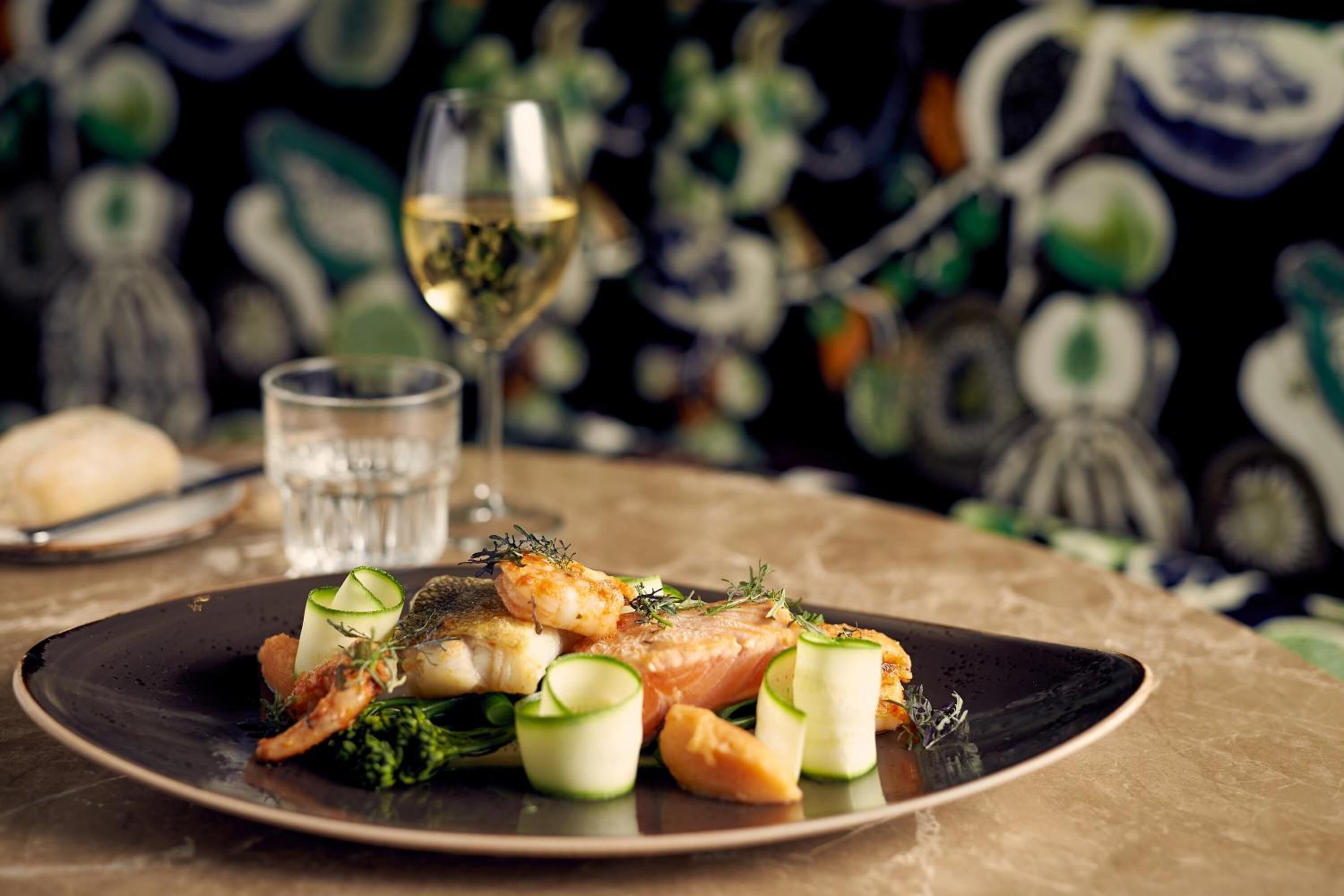 Une assiette de nourriture, un verre de vin blanc et un petit verre d’eau sont posés sur une table.