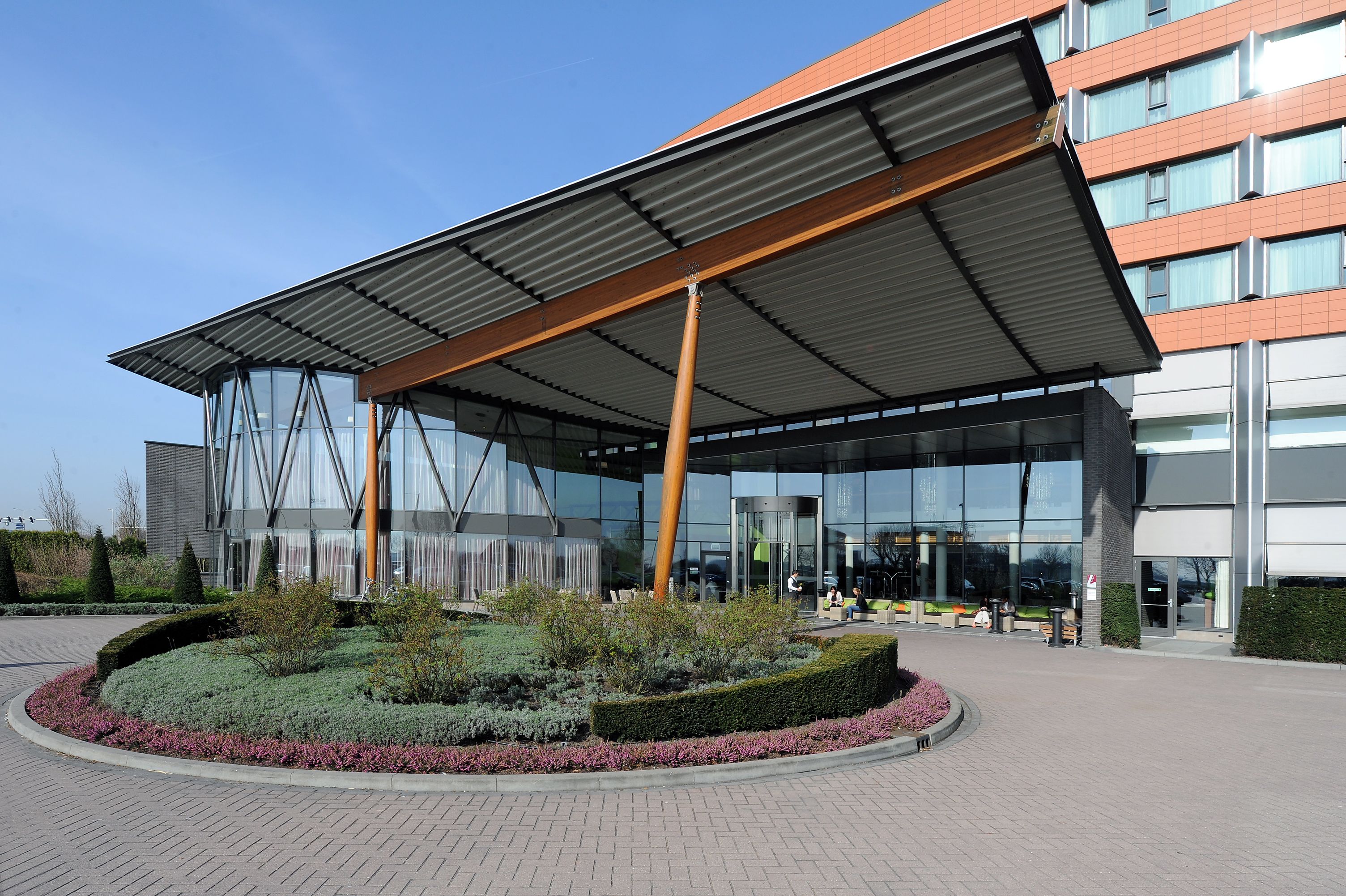 Een modern gebouw met een glazen gevel, een overdekte ingang en aangelegde tuinen.