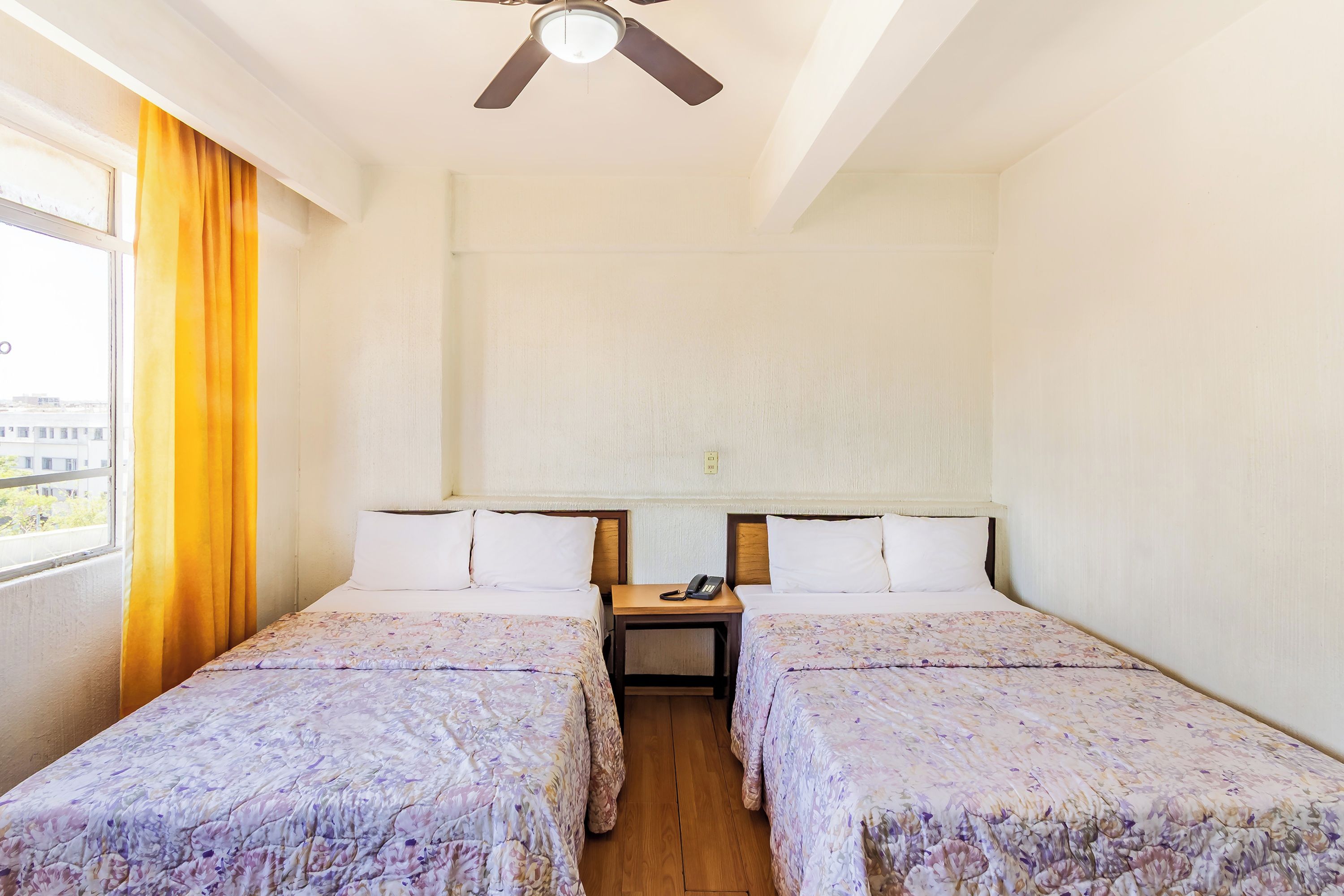 A hotel room with two beds, a ceiling fan, a nightstand with a phone, and a window with curtains.