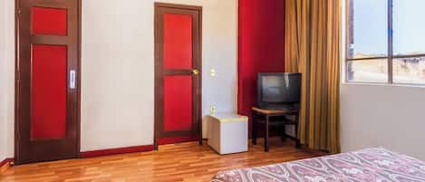 A hotel room with wooden flooring, two red doors, a bed with a patterned bedspread, a television on a small table, and a window with curtains.