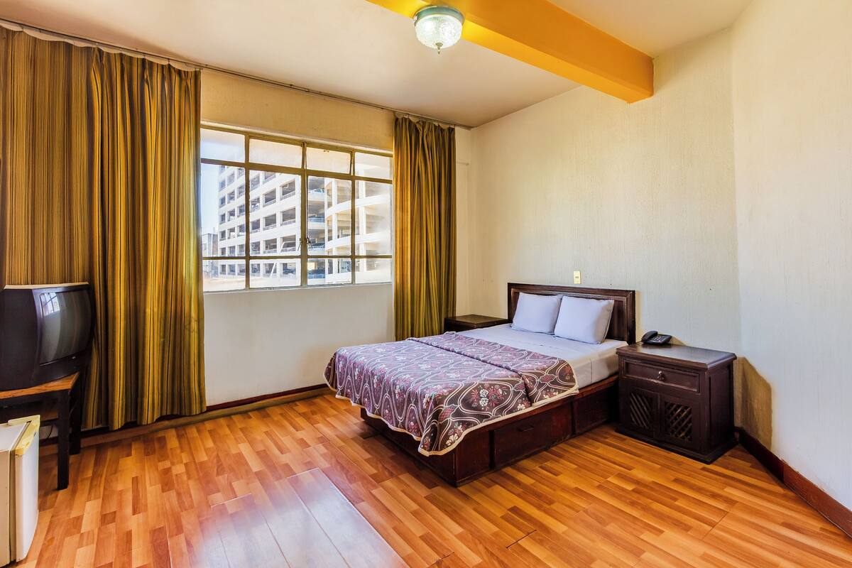 A hotel room with a wooden floor, a bed with a patterned bedspread, a bedside table with a phone, and a television on a stand.
