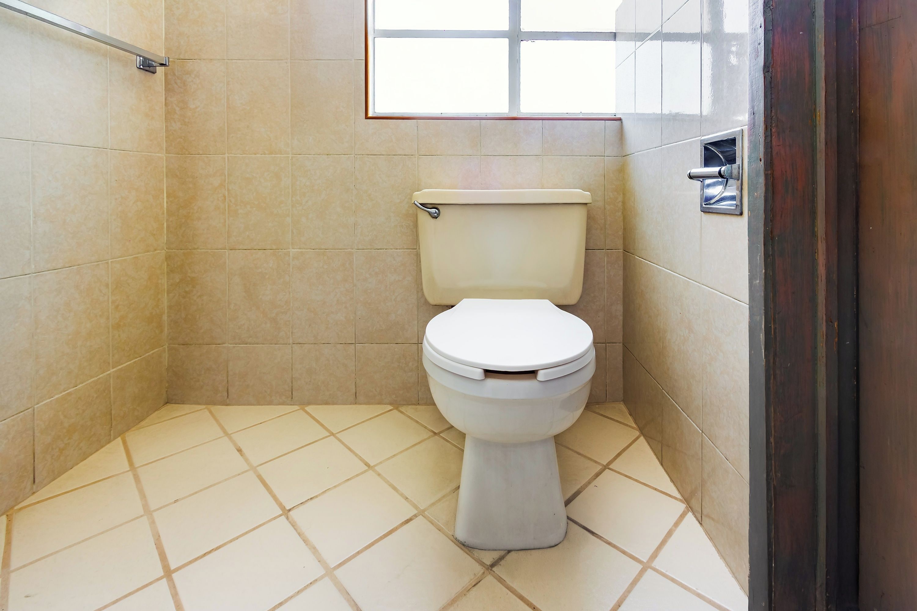 A small, tiled bathroom with a toilet, a window, and a metal handrail.