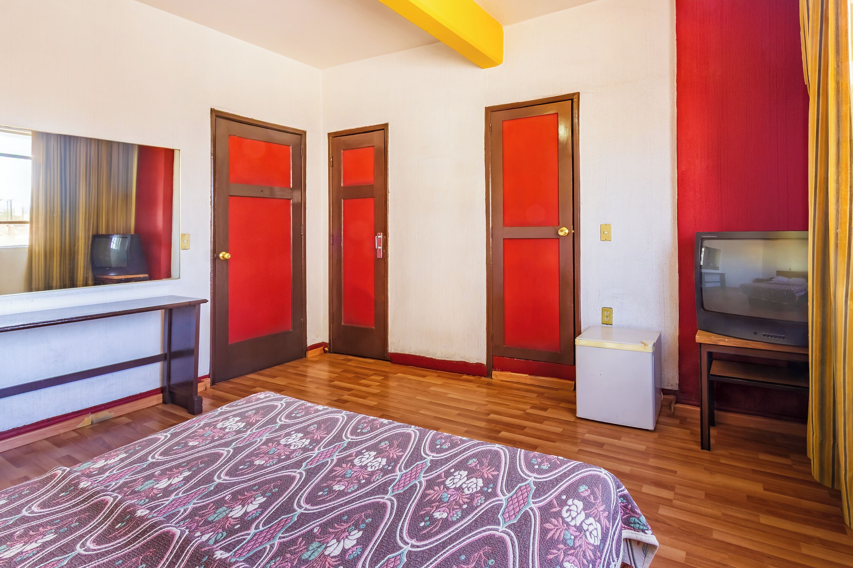 Una camera d'albergo con pavimento in legno, un letto con copripiumino a motivi floreali, una televisione su un mobile e tre porte con pannelli rossi.