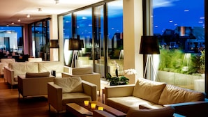 A modern hotel lobby with leather sofas, a wooden coffee table, and floor lamps. Large windows offer a view of the cityscape at night. - Midori The Green Guesthouse (Dossenheim)