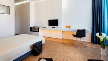 Una habitación de hotel moderna con cama, escritorio con televisor de pantalla plana, silla, jarra con flores y un bolso en el suelo.