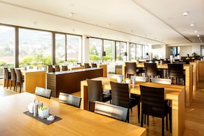 Een ruime restaurantzaal met meerdere eettafels en stoelen, grote ramen met uitzicht op het omliggende landschap, en een modern interieur.
