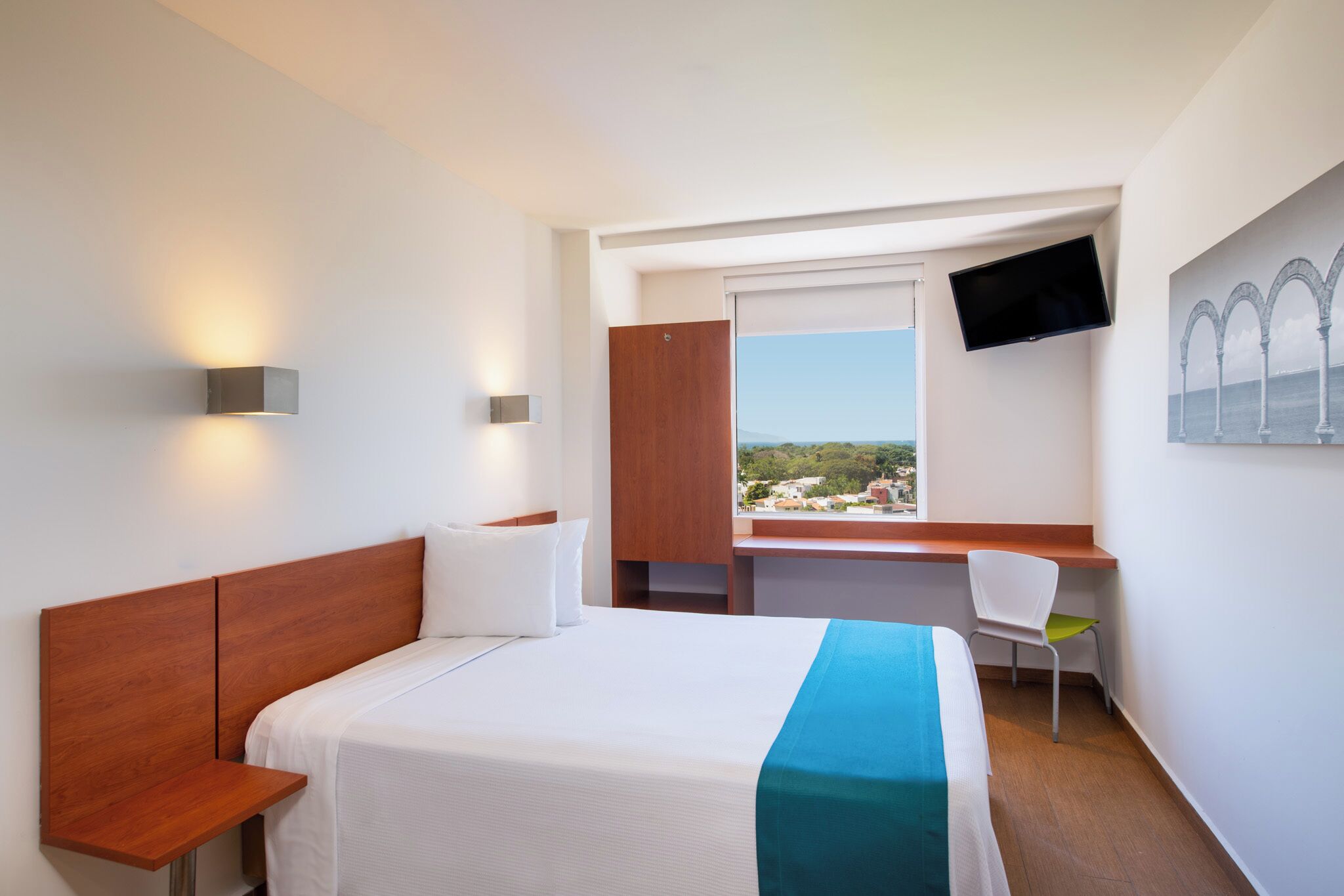 A hotel room with a bed, a desk, a chair, a TV, and a window with a view.