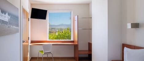 A hotel room with a bed, a desk with a chair, a TV mounted on the wall, and a window with a view of greenery and mountains.